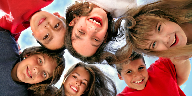 Glada skolbarn står i ring lutad över kameran med en blå himmel som bakgrund