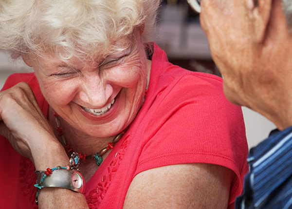 Äldre kvinna i röd blus sitter vid ett bord och skrattar tillsammans med en äldre man i blå skjorta