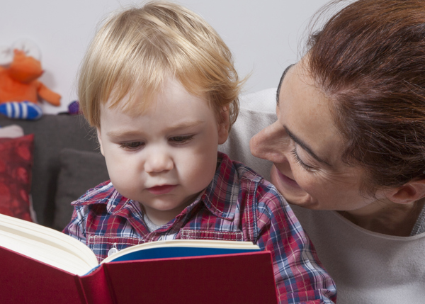 Kvinna sitter på golvet med ett litet barn i knäet och läser ur en röd bok